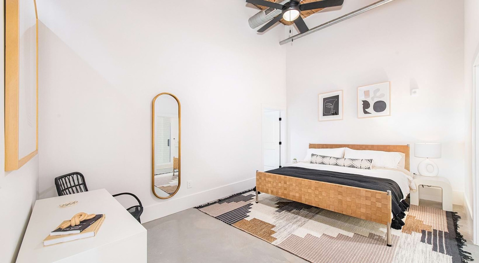 A bedroom with a woven leather bed frame, a graphic rug, an oval gold mirror, a white desk, and a ceiling fan below an exposed duct