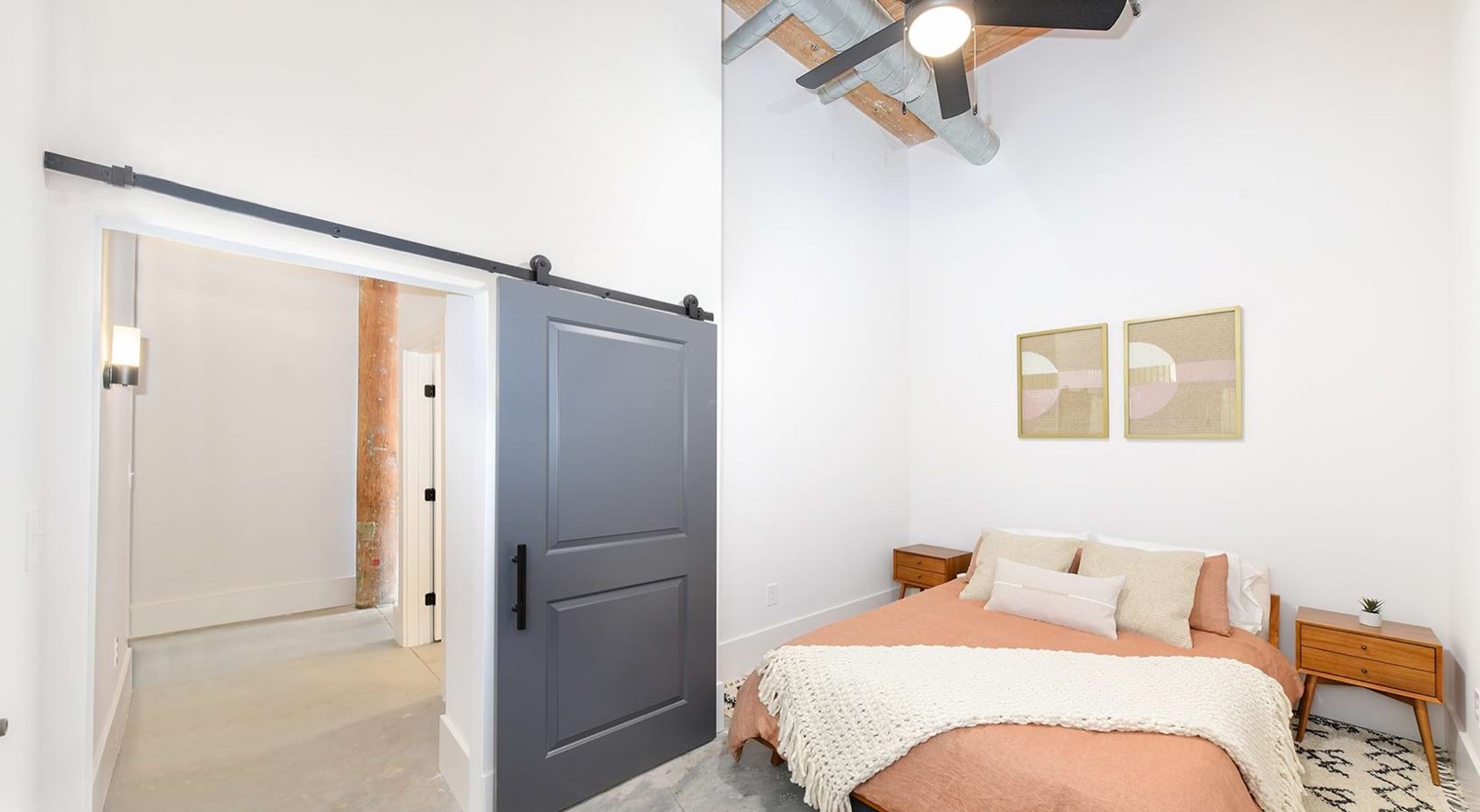 A bedroom with an orange linen bed, walnut nightstands, a gray barn door, and a ceiling fan beneath exposed timber and ductwork