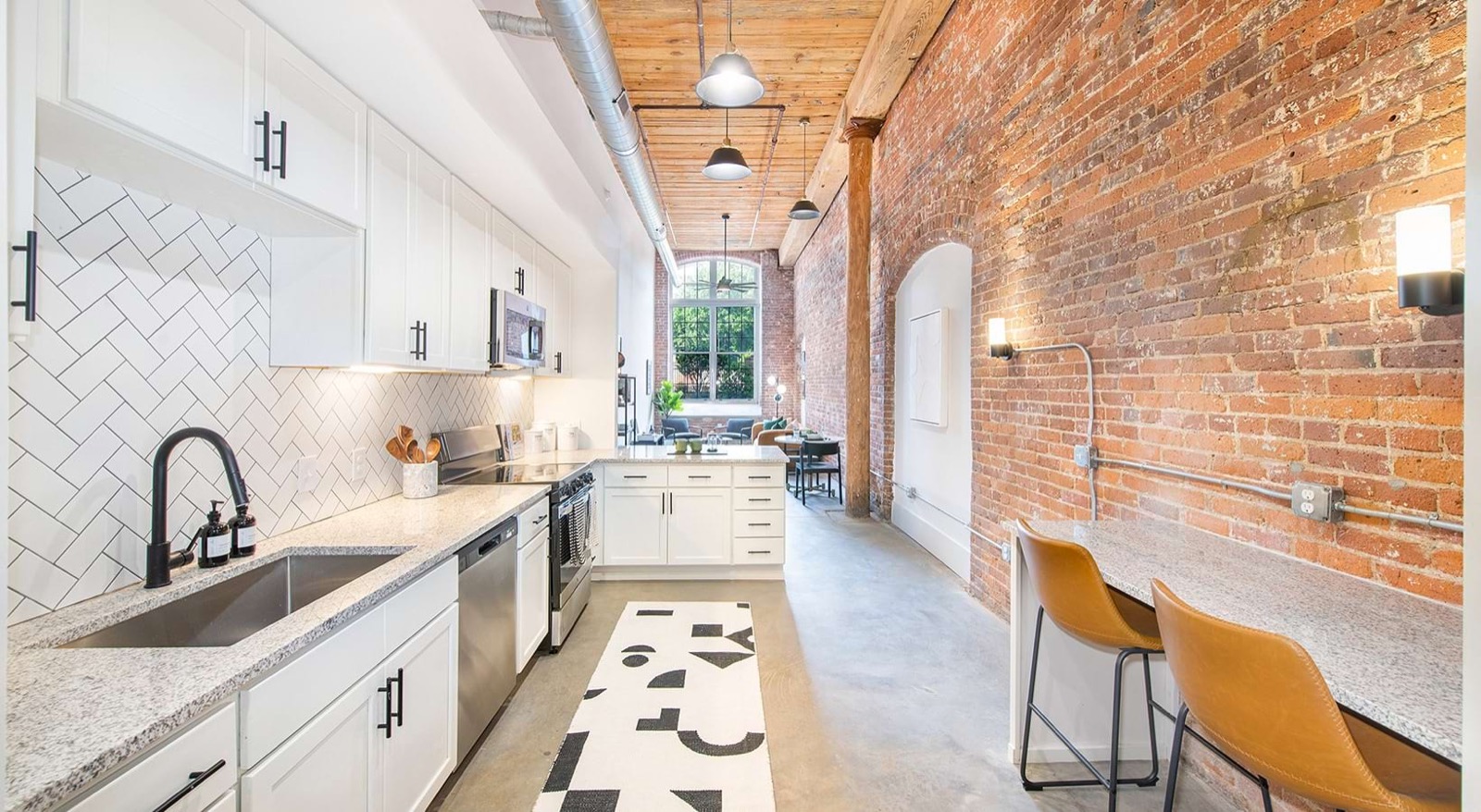 A long galley kitchen with white cabinets, a herringbone tile backsplash, granite counters, and tan leather stools beside an exposed brick wall
