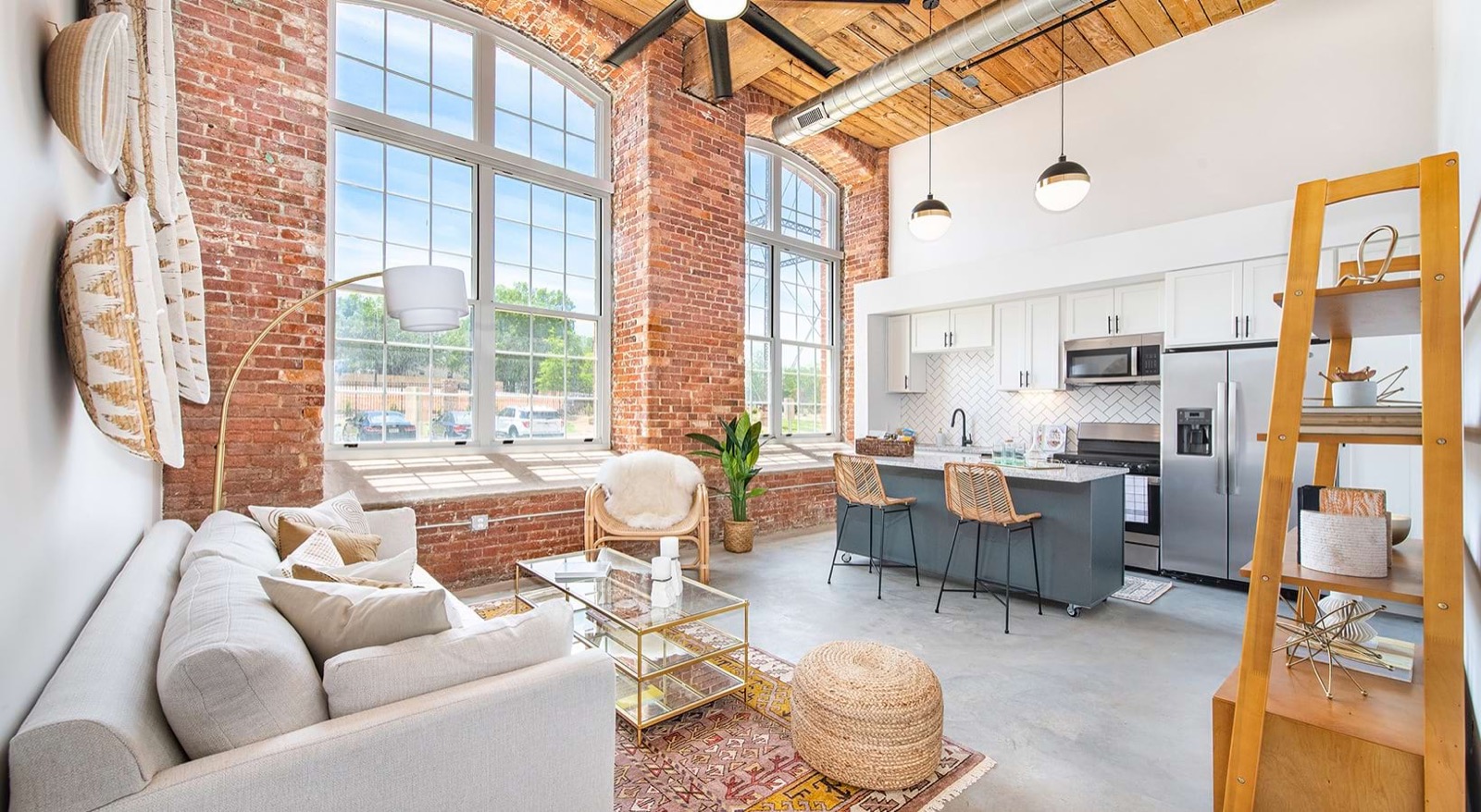 A loft living room with tall arched windows, a cream sofa, a rattan chair, a gold coffee table, and a kitchen island in dark gray