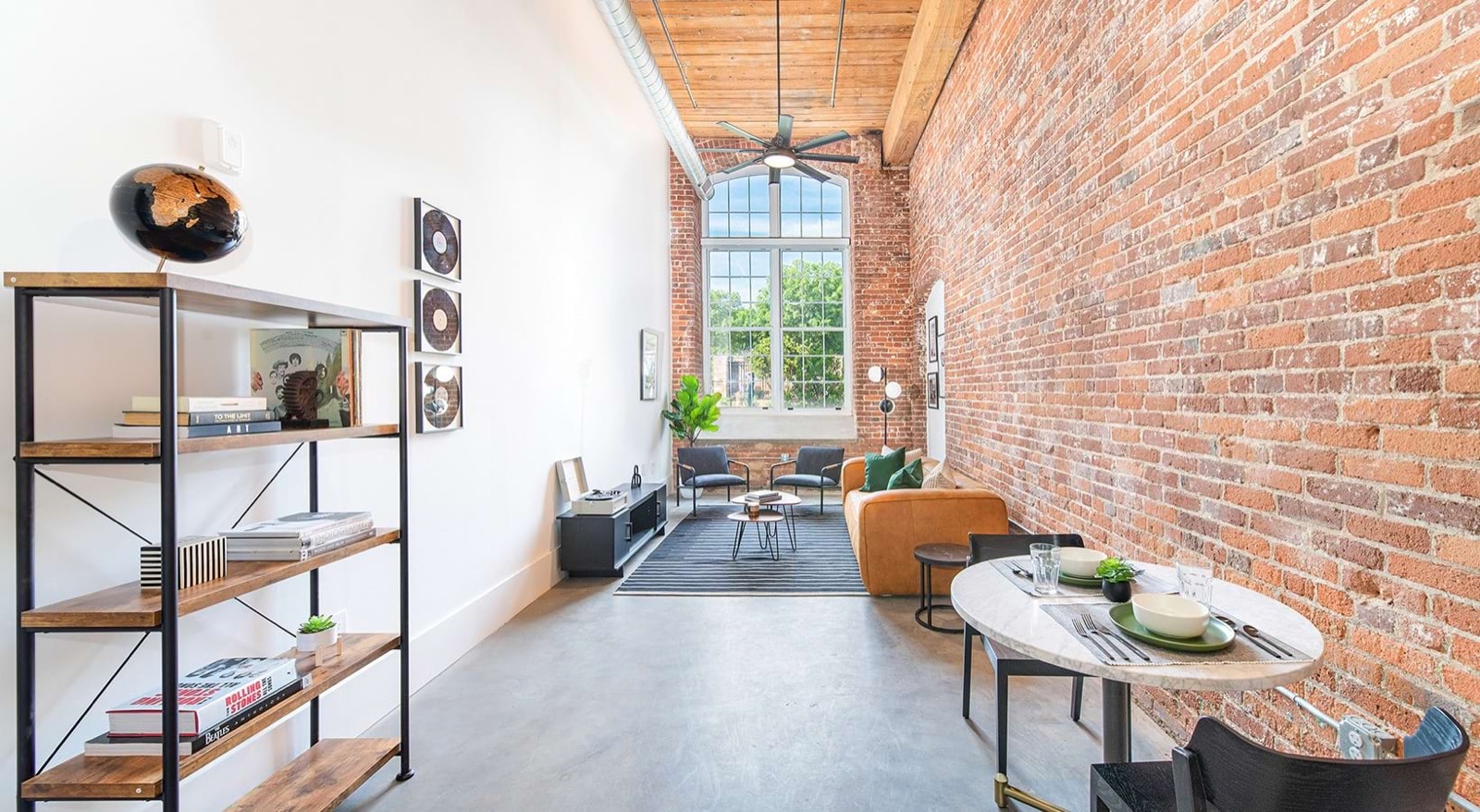 A narrow living room with an exposed brick wall, a tan leather sofa, a striped rug, an iron shelf with books and a globe, and a round marble dining table