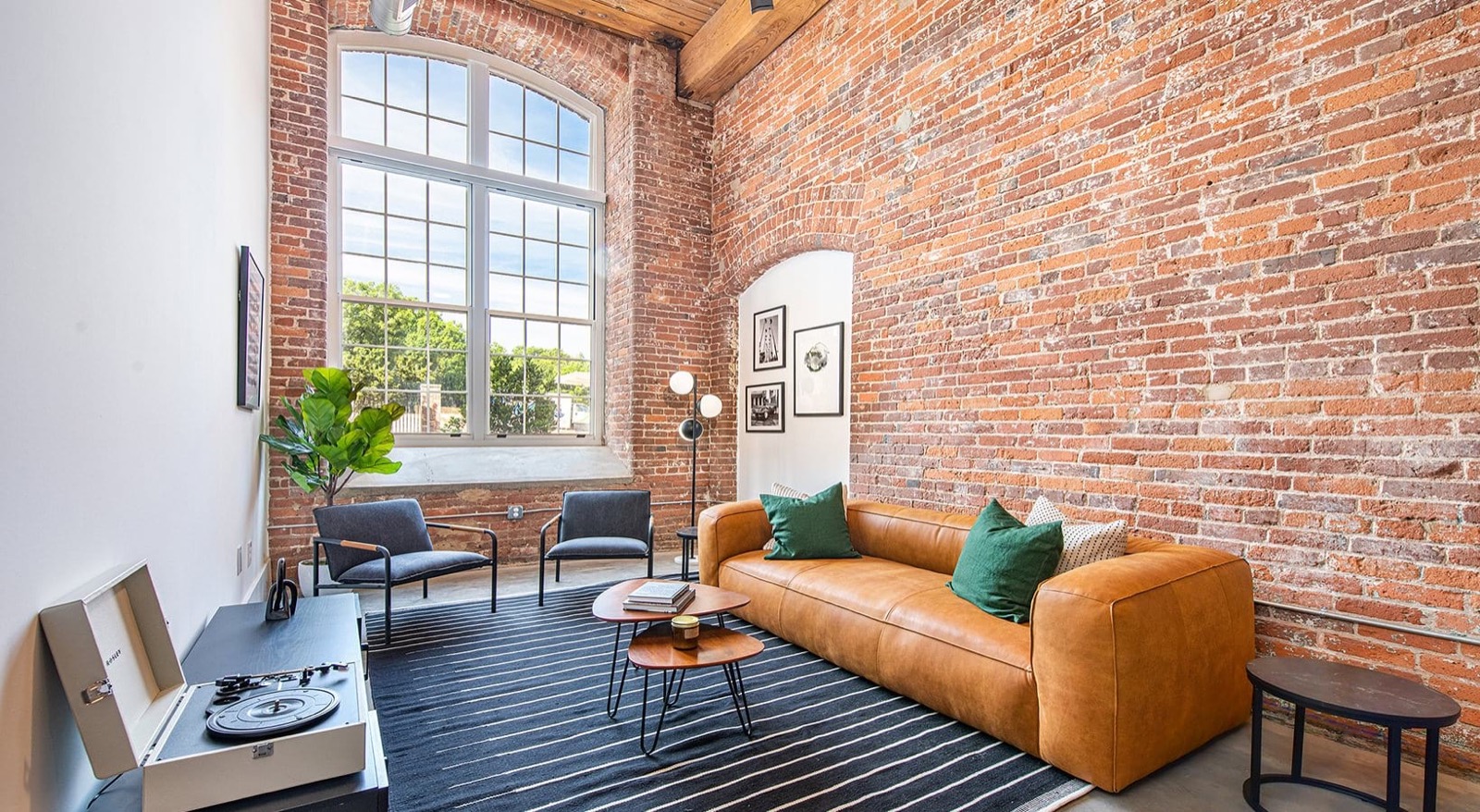 A living room corner with a tan leather sofa, gray armchairs, a record player, and an arched doorway in an exposed brick wall