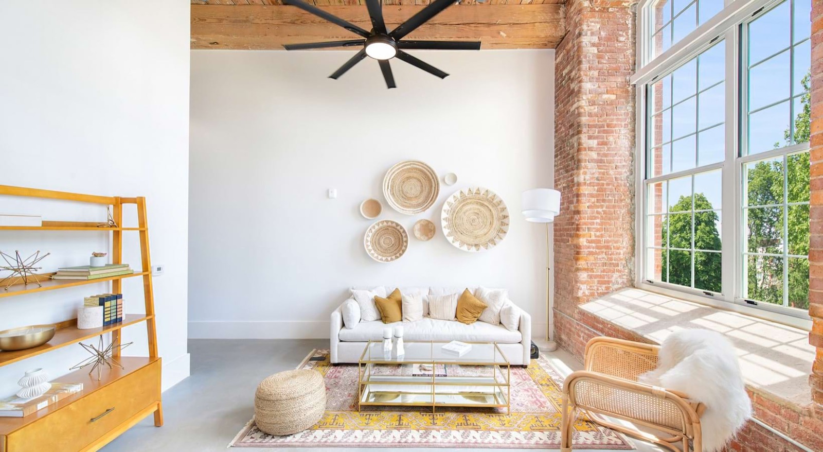 A white living room with a cream sofa, a gold coffee table, a ring of woven baskets on the wall, an amber shelf, and a ceiling fan