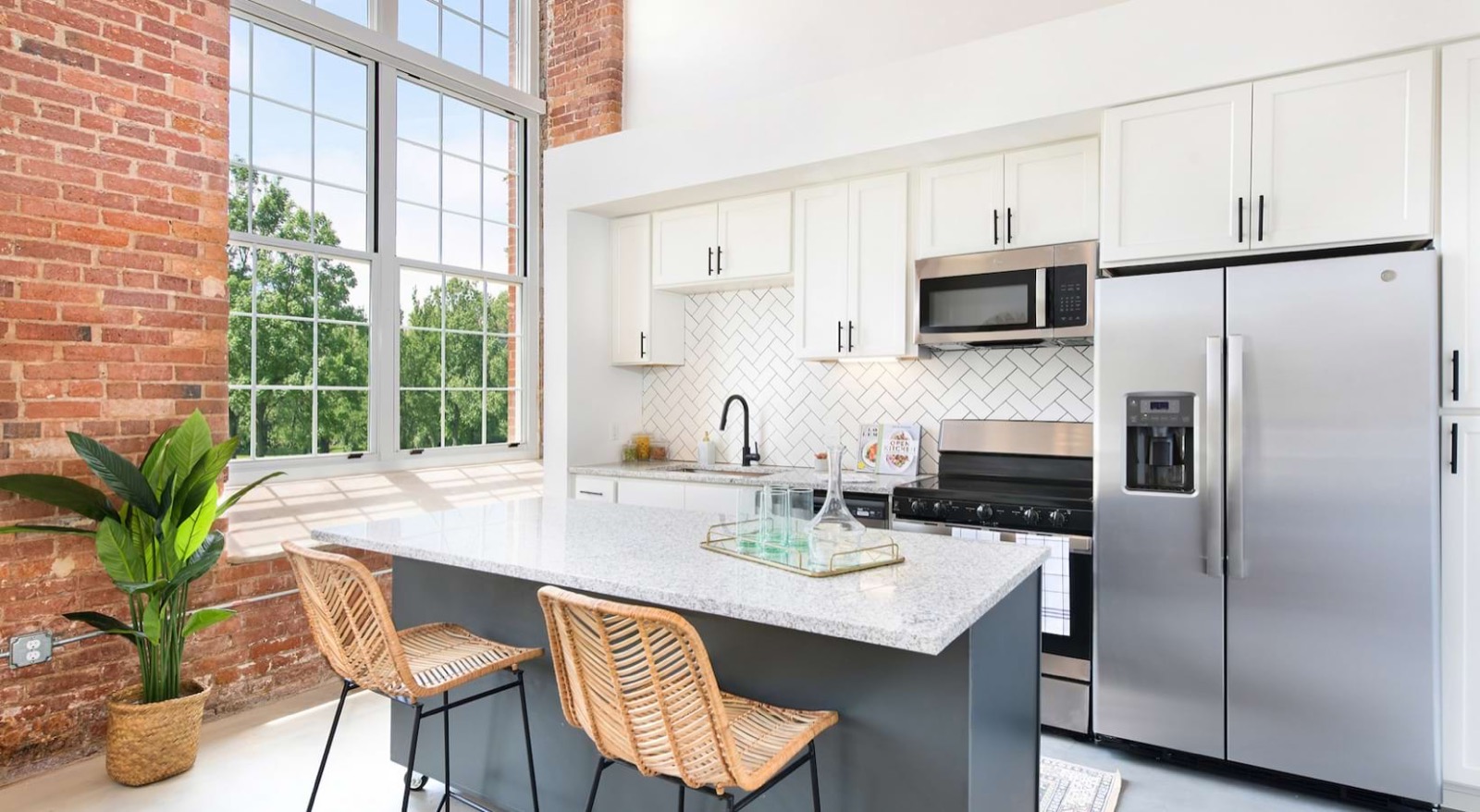A kitchen with white cabinets, a herringbone backsplash, a dark gray island with granite and rattan stools, and a stainless refrigerator