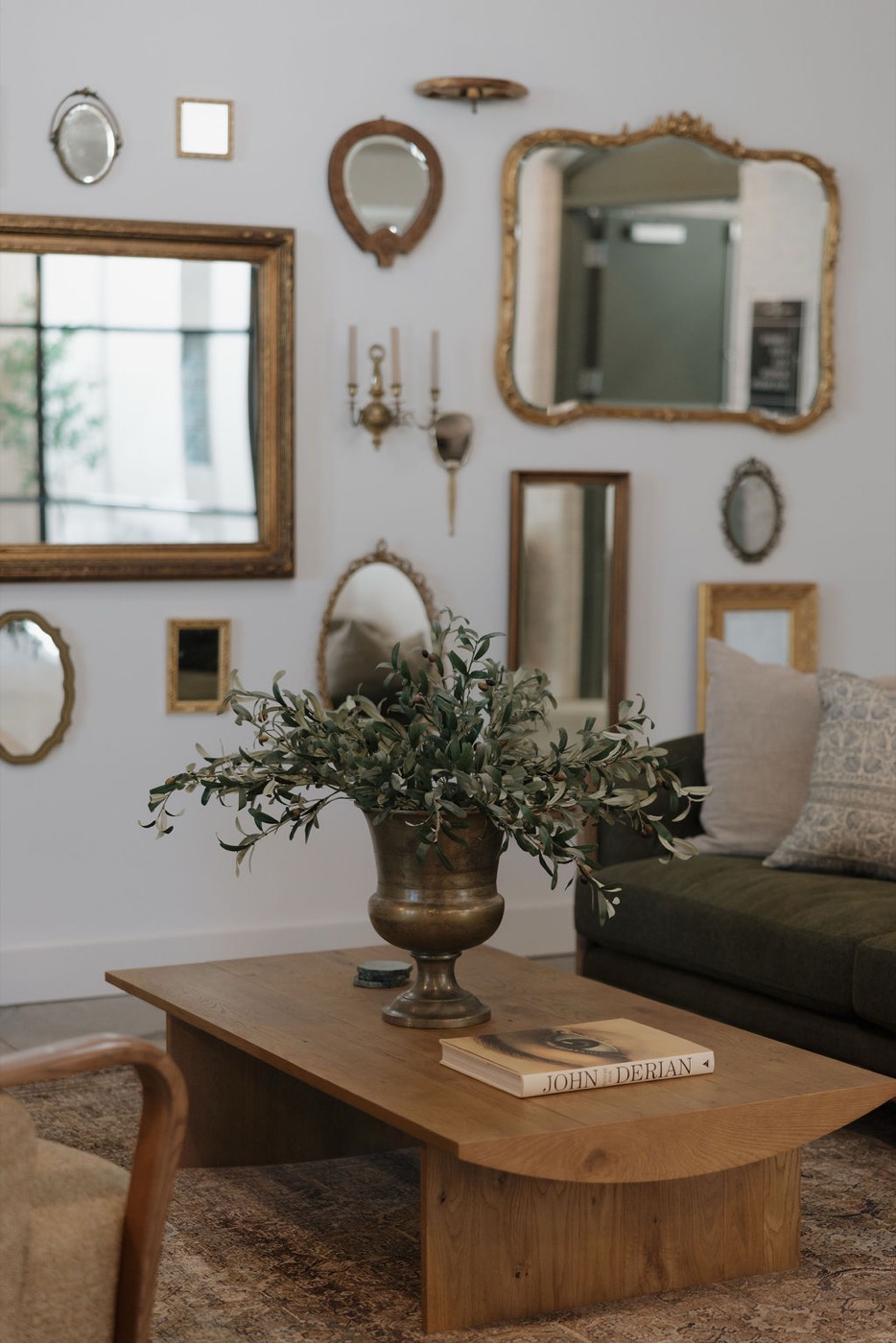 Gallery wall of antique gilded mirrors above a wood coffee table with a brass urn of olive branches and a green velvet sofa